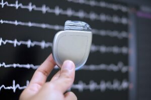 Hand holding a cardiac pacemaker device in front of an ECG monitor displaying heart rhythm data